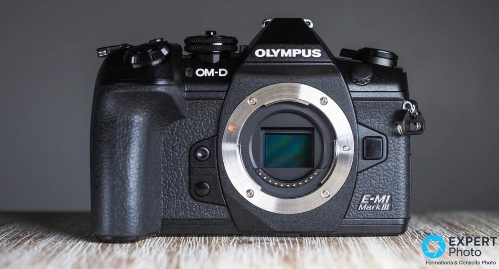Olympus OM-D E-M1 Mark III de face, bouchon retiré, monture Micro 4/3 et capteur visibles sur fond gris studio. 