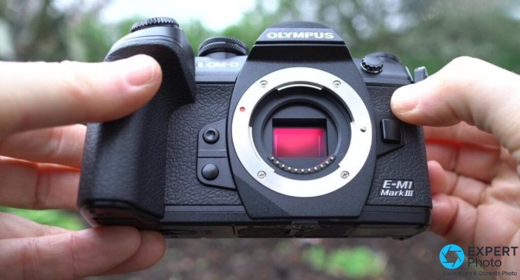 Olympus OM-D E-M1 Mark III tenu à deux mains face avant, bouchon retiré, capteur visible, extérieur vert flou.