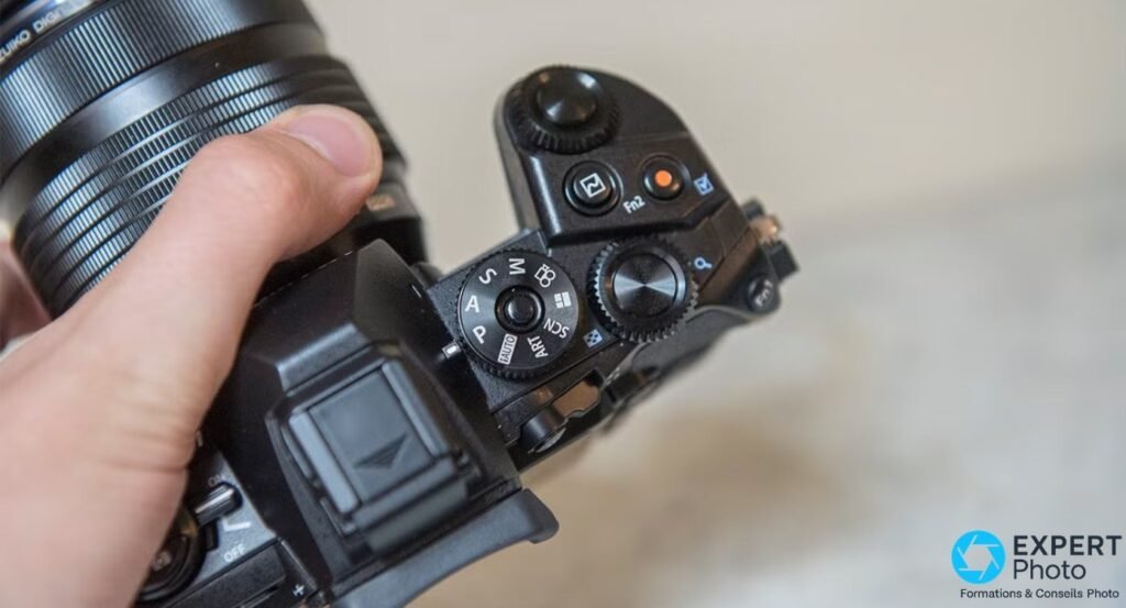 Olympus OM-D E-M1 en main, vue de dessus sur molette PASM, boutons Fn2 et déclencheur, objectif M.Zuiko visible. 