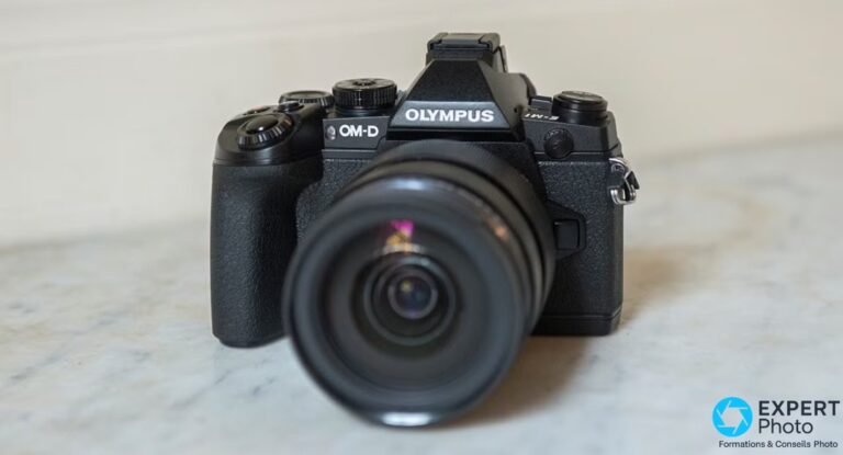 Boîtier Olympus OM-D E-M1 noir vu de 3/4 avant avec objectif monté, posé sur surface marbrée claire.