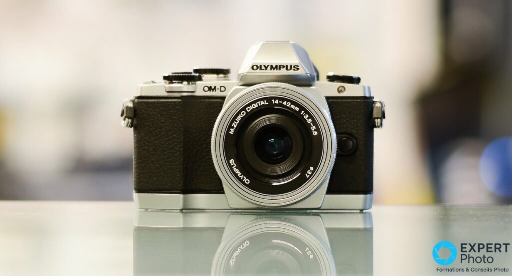 Olympus OM-D E-M10 coloris argent de face avec zoom M.Zuiko Digital 14-42 mm posé sur surface réfléchissante