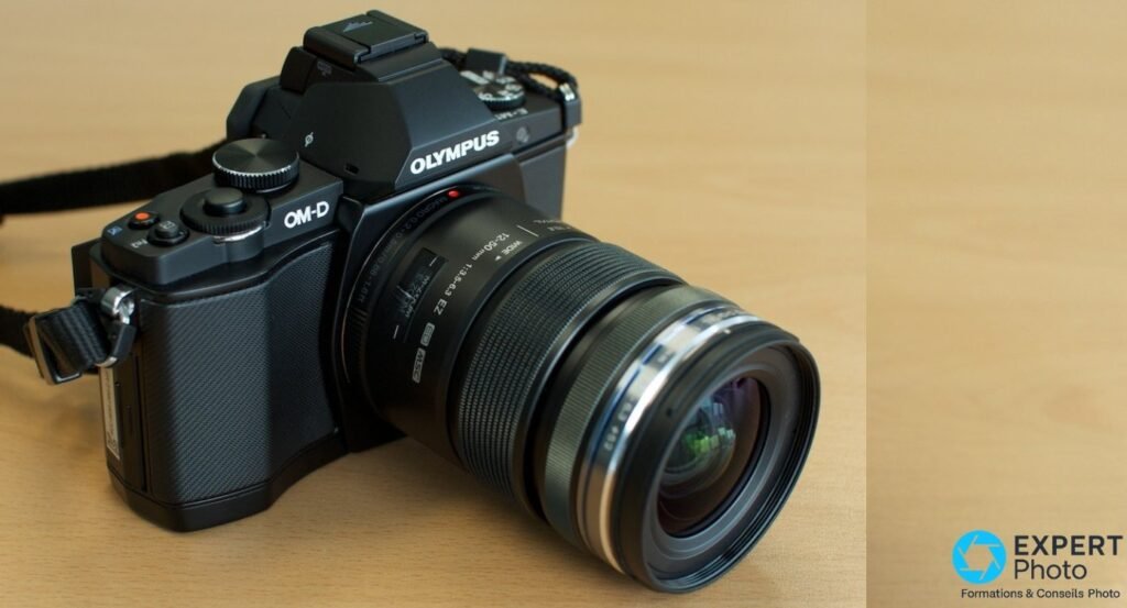 Olympus OM-D E-M5 noir avec zoom M.Zuiko 12-50 mm monté, vu de 3/4 avant, posé sur bois clair.
