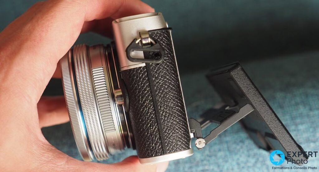 Olympus PEN E-P7 de profil tenu à la main, objectif M.Zuiko monté, écran tactile rabattu à 180° en position selfie.