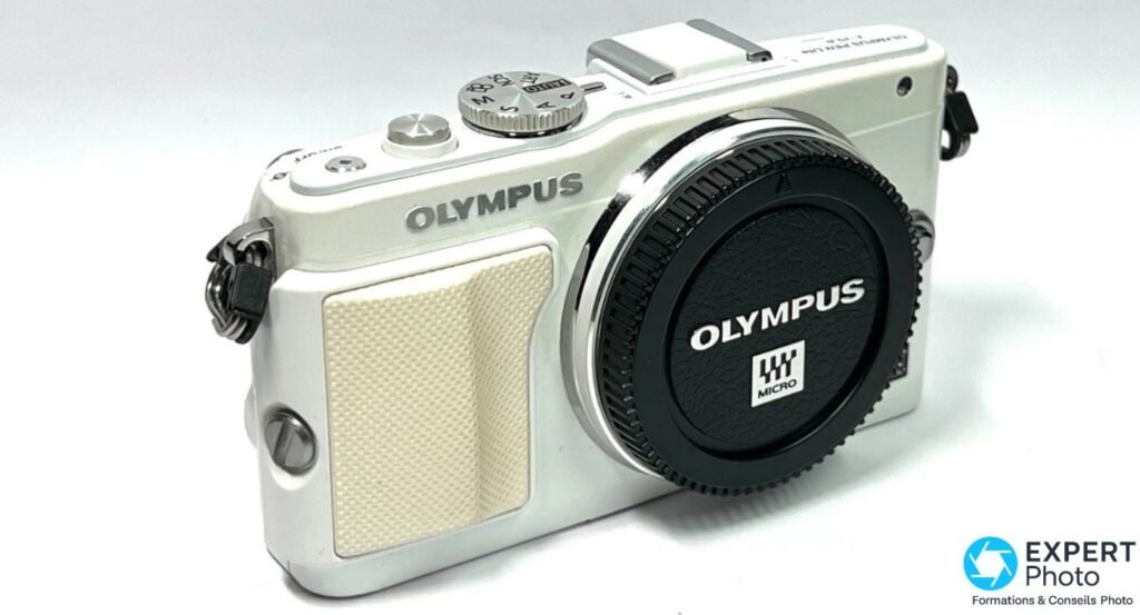 Olympus PEN E-PL6 blanc vu de face en trois quarts, cache de monture Micro Four Thirds en place, logo OLYMPUS sur le front