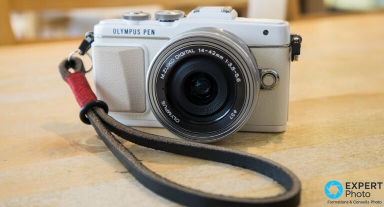Olympus PEN E-PL7 blanc vu de 3/4 avant, objectif 14-42mm monté, dragonne cuir noire et rouge, posé sur table bois