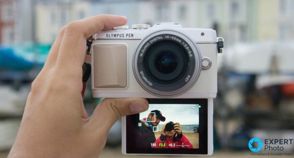 Olympus PEN E-PL7 blanc tenu en main droite, écran LCD basculable déplié vers le bas, arrière-plan de port flou 