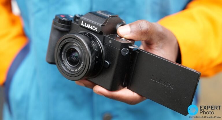 Test Panasonic Lumix G100 : l’hybride vlog à viseur, pour un profil précis Photographe tenant le Panasonic Lumix G100 à deux mains face à lui, logo LUMIX visible au-dessus du capteur, mains baguées