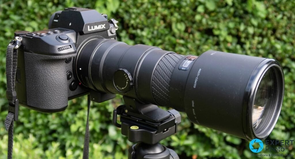 Boîtier Panasonic Lumix S avec téléobjectif noir sur embase trépied devant haie de feuillage vert.