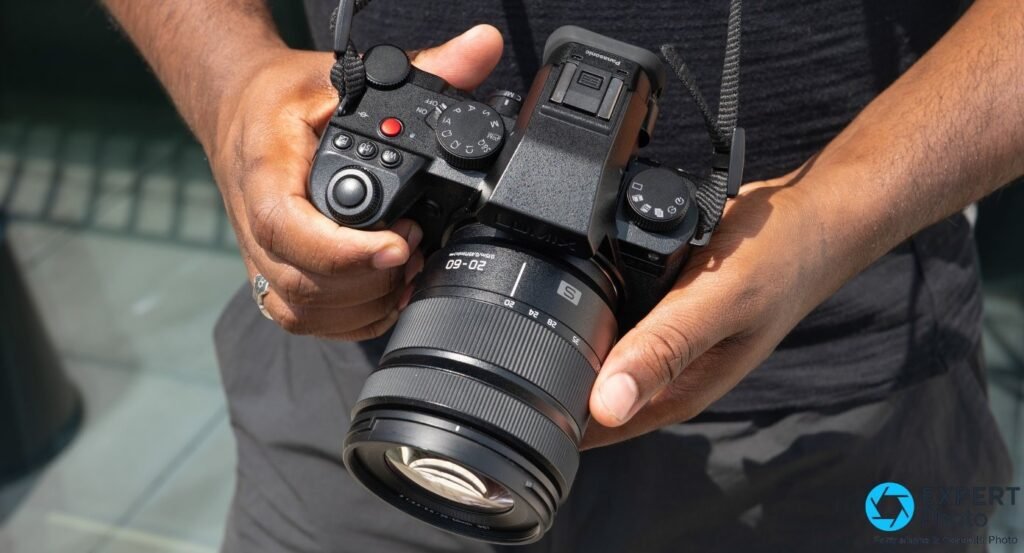  Panasonic Lumix S5 IIX tenu en main, vue du dessus, zoom Lumix S 20-60mm monté, molettes de modes et déclencheur visibles.