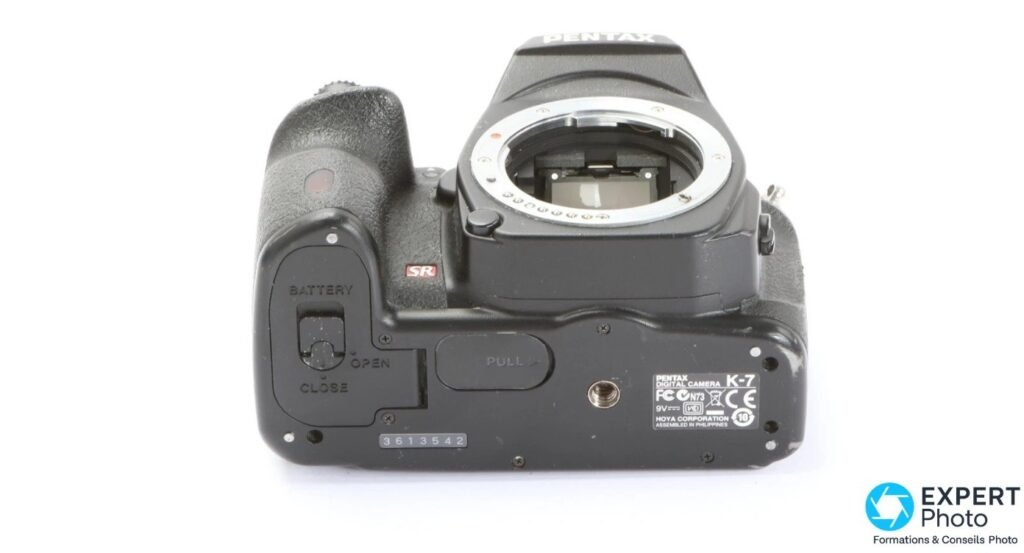 Dessous du Pentax K-7, trappe batterie, pas de vis trépied et plaque signalétique indiquant le numéro de série. 