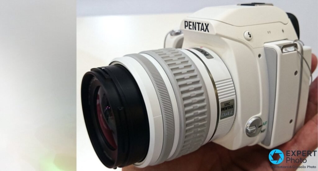 Pentax K-S1 blanc vu de 3/4 côté gauche, zoom SMC Pentax DAL monté bouchon avant noir en place, main partielle à droite. 