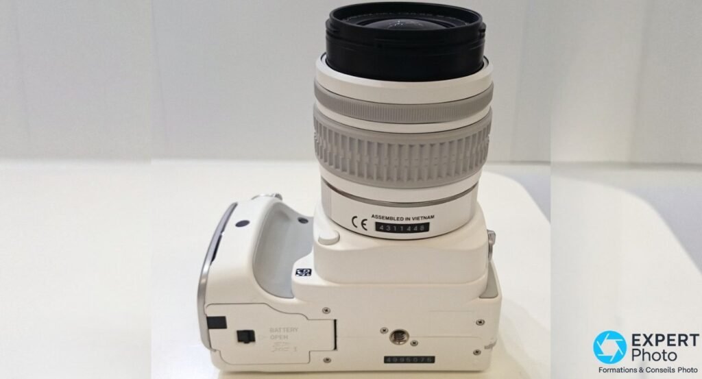  Dessous du boîtier Pentax K-S1 blanc, trappe batterie SDXC fermée et filetage trépied, zoom SMC Pentax DAL monté. 