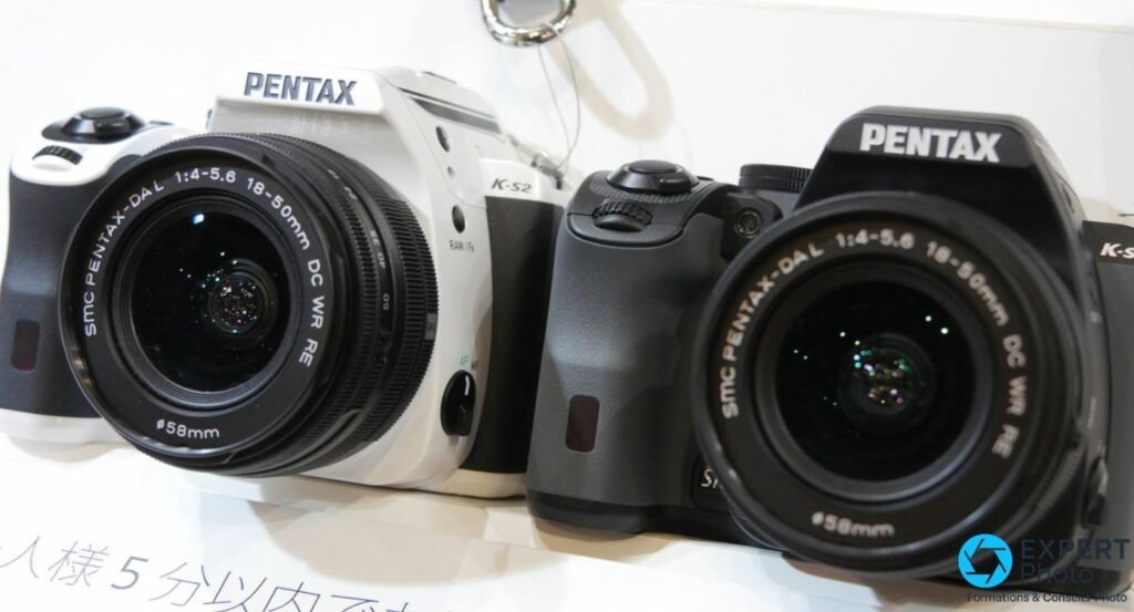 Pentax K-S2 noir équipé du zoom SMC Pentax-DAL 18-50mm f/4-5.6 DC WR RE, vue face trois quarts sur plateau d'exposition