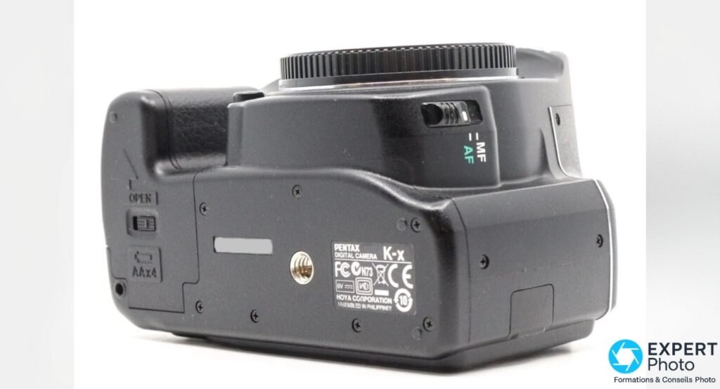 Pentax K-x de dessous, plaque HOYA Corporation, trappe OPEN marquée AAx4 pour 4 piles AA et filetage trépied. 