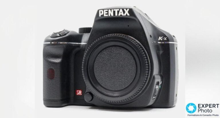 Pentax K-x de face, boîtier reflex APS-C noir, logos PENTAX et K-x visibles, cache de boîtier monté, marquage SR rouge.