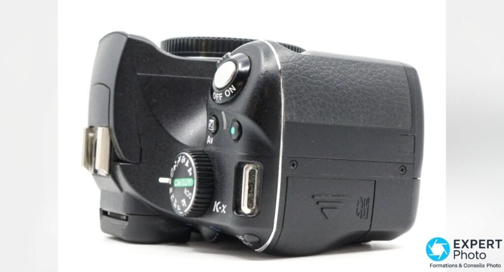 Pentax K-x vu du dessus en gros plan, molette des modes SCN et cache d'un port de connexion ouvert sur le côté droit. 