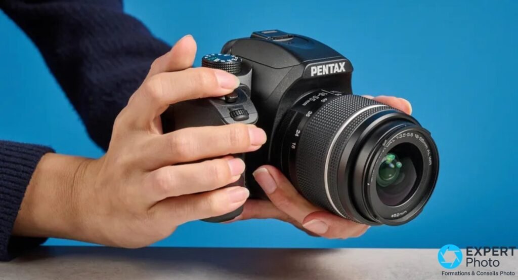 Pentax KF avec objectif 18-55 WR tenu à deux mains en position de prise de vue, studio fond bleu