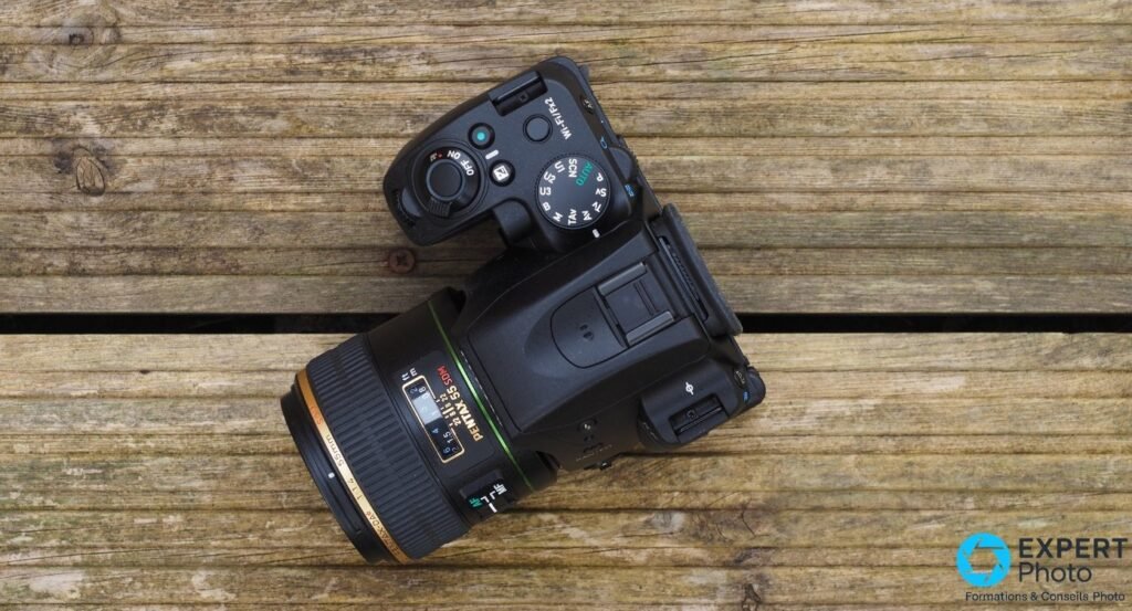 Pentax KF en vue plongée avec objectif Pentax 55 mm f/1.4 SDM, échelle de diaphragme visible, sur planches de bois 