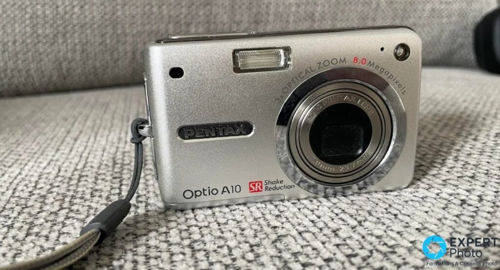 Pentax Optio A10 argenté en 3/4 avant avec dragonne noire, posé sur un canapé tissu gris à chevrons. 