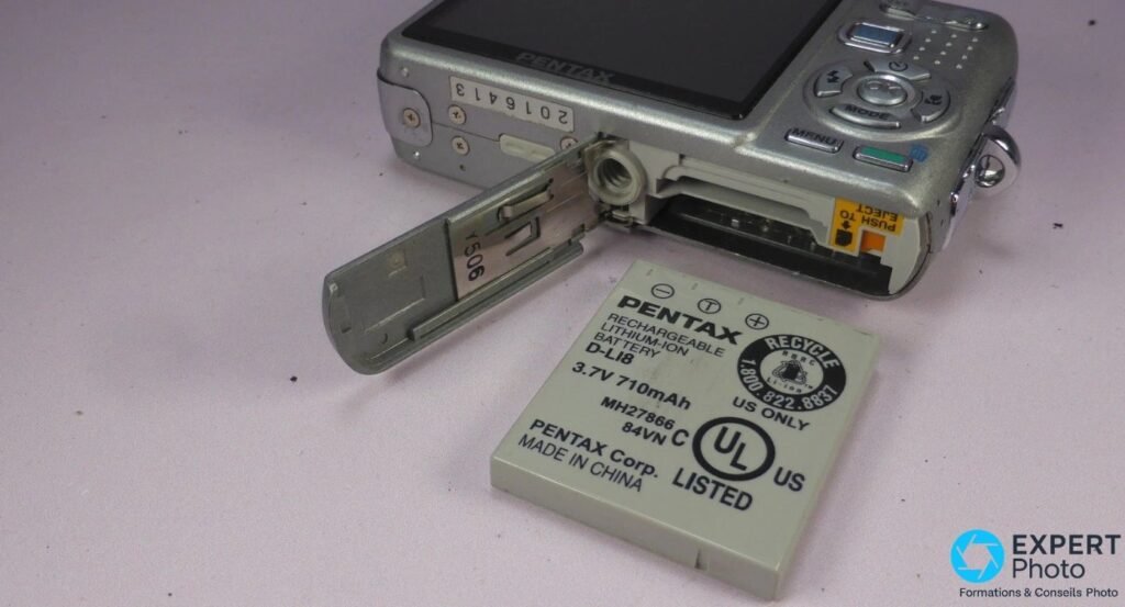 Pentax Optio A20 silver, trappe batterie ouverte, batterie Pentax D-LI8 3,7V 710mAh posée à côté.