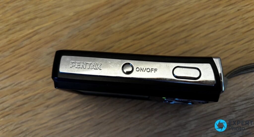 Pentax Optio E60 vu du dessus, bouton ON/OFF central, déclencheur à droite et logo Pentax gravé à gauche