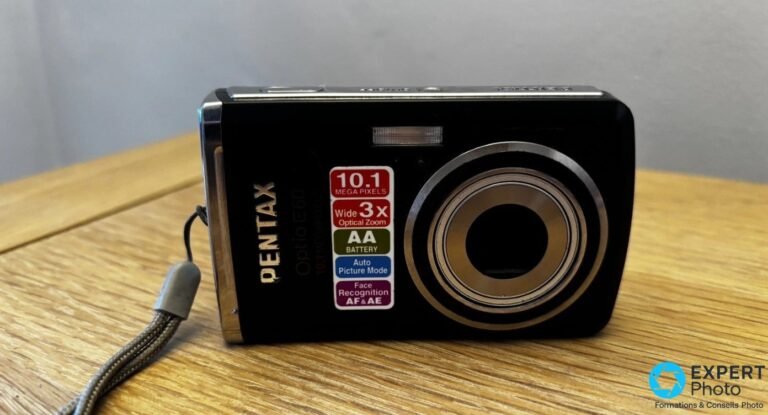 Pentax Optio E60 noir posé sur table, face avant, objectif rétracté, étiquettes 10,1 Mpx, zoom 3x et piles AA visibles