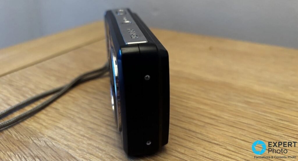 Pentax Optio E60 noir vu de profil droit, logo Pentax gravé sur la tranche supérieure, dragonne posée sur la table