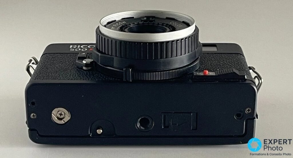 Semelle du Ricoh 500 ME avec pas de vis trépied au centre, bouton de rembobinage, objectif pointant vers le haut. 