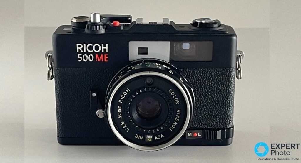 Face avant frontale du Ricoh 500 ME, objectif Color Rikenon 40 mm centré, curseur M-E et logo RICOH 500 ME visibles. 