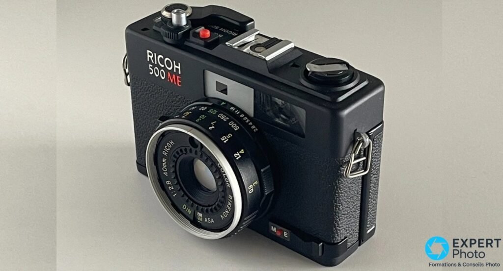 Ricoh 500 ME, trois-quarts avant côté droit, curseur M-E rouge du sélecteur manuel-automatique visible sous l objectif. 
