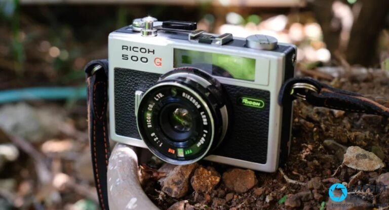 Ricoh 500G vu trois-quarts avant, boîtier ivoire cuir noir, objectif Rikenon 40 mm f/2,8 sur fond nature flou.
