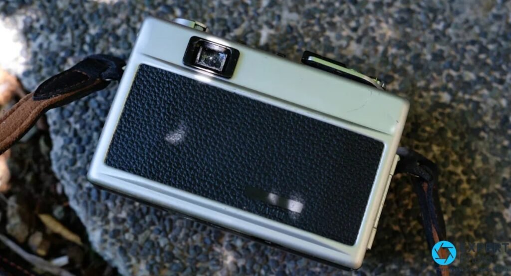 Dos du Ricoh 500G ivoire et cuir noir avec sangle en cuir tressée noir et marron, posé sur graviers.