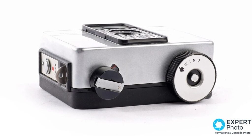 Ricoh Auto Half E en vue trois-quarts arrière sur fond blanc, grosse molette WIND argentée de remontage visible. 