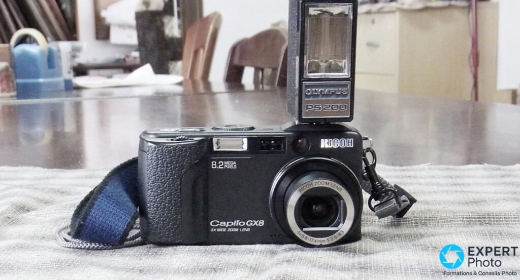 Ricoh Caplio GX8 vu de face avec un flash externe Olympus PS200 monté sur la griffe, sangle bleue visible. 