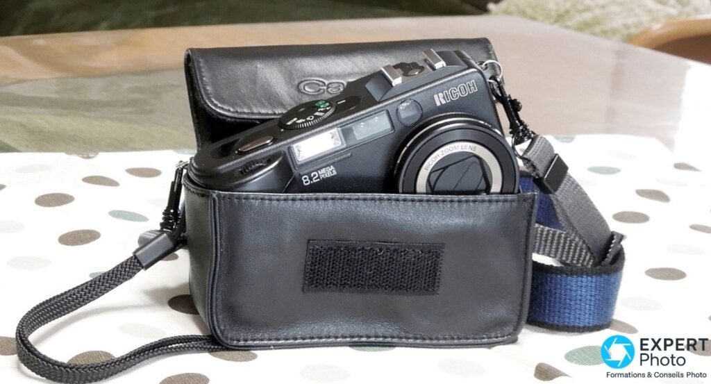 Ricoh Caplio GX8 partiellement sorti de son étui en cuir noir, posé sur une nappe à pois beiges, sangle bleue visible.