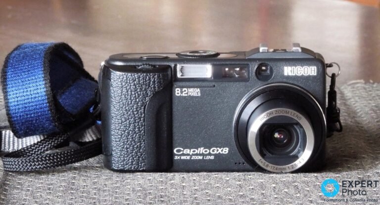 Ricoh Caplio GX8 noir vu de face avec sa sangle bleue et grise, zoom Ricoh 5,8-17,4 mm rétracté, fond gris uni.