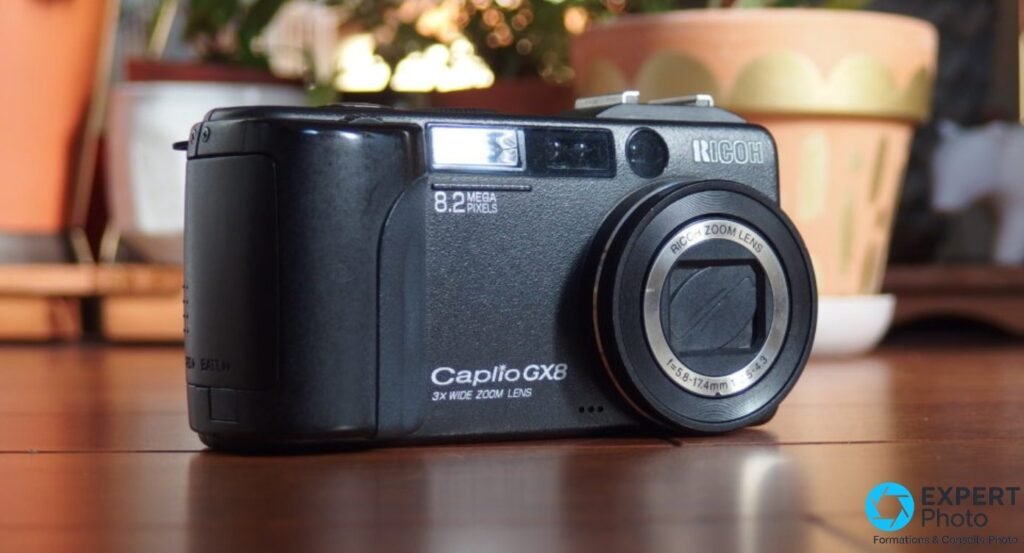 Ricoh Caplio GX8 noir vu de trois quarts droit, zoom Ricoh 5,8-17,4 mm rétracté, plantes en arrière-plan flou.