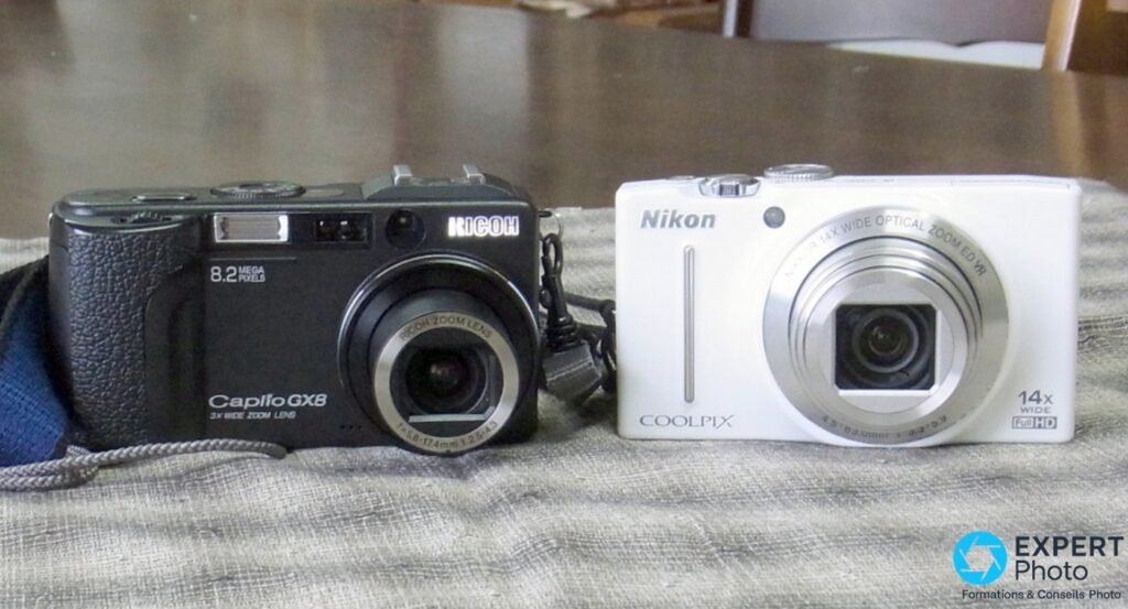 Ricoh Caplio GX8 noir et compact Nikon Coolpix blanc 14x posés côte à côte sur un tissu rayé clair.
