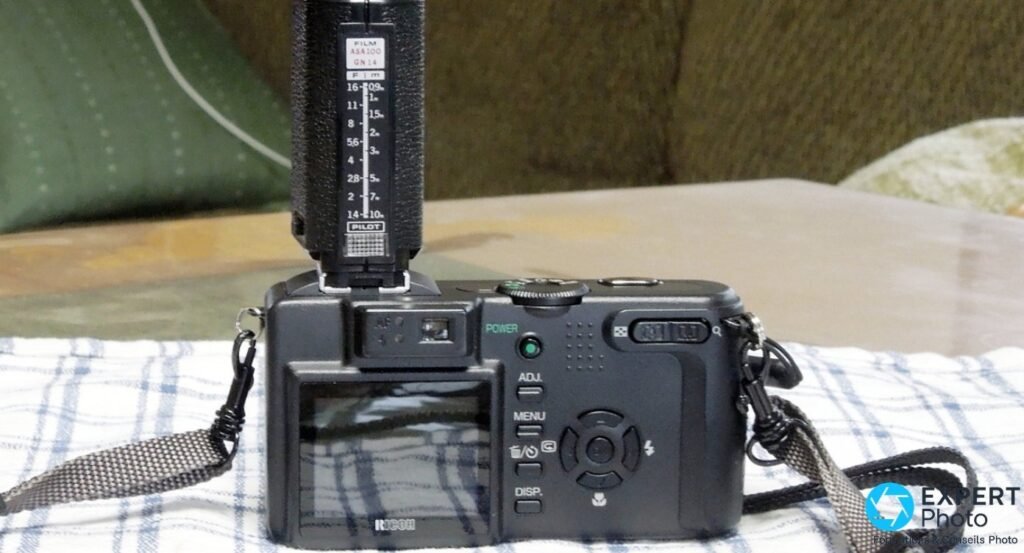 Dos du Ricoh Caplio GX8 montrant l'écran LCD, les boutons MENU, ADJ et DISP, avec un flash externe à échelle ASA monté.