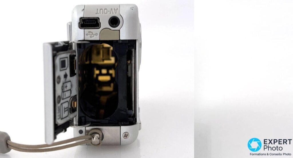 Compartiment batterie du Ricoh Caplio R2 ouvert montrant contacts dorés, fente carte SD et ports USB AV-OUT au-dessus. 