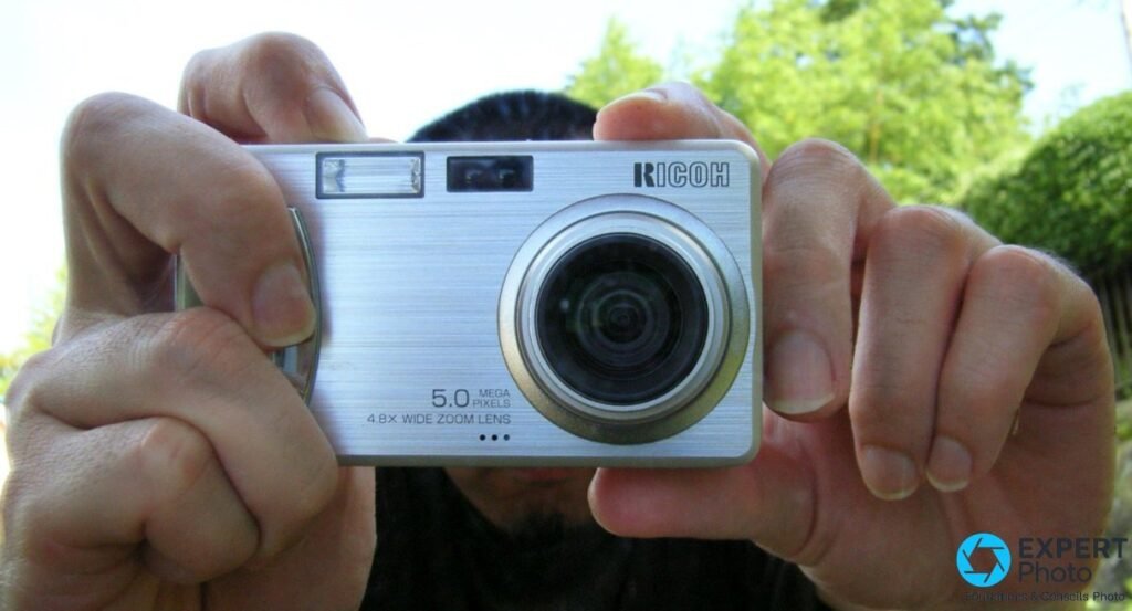 Photographe en extérieur tenant le Ricoh Caplio R2 à deux mains à bout de bras, devant un fond d'arbres verts. 