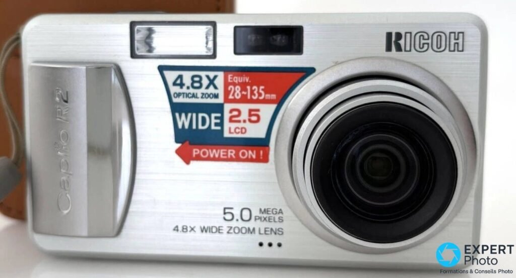 Ricoh Caplio R2 argenté vu de face, sticker d'origine 4,8x optical zoom 28-135 mm visible, étui cuir flou en arrière-plan.