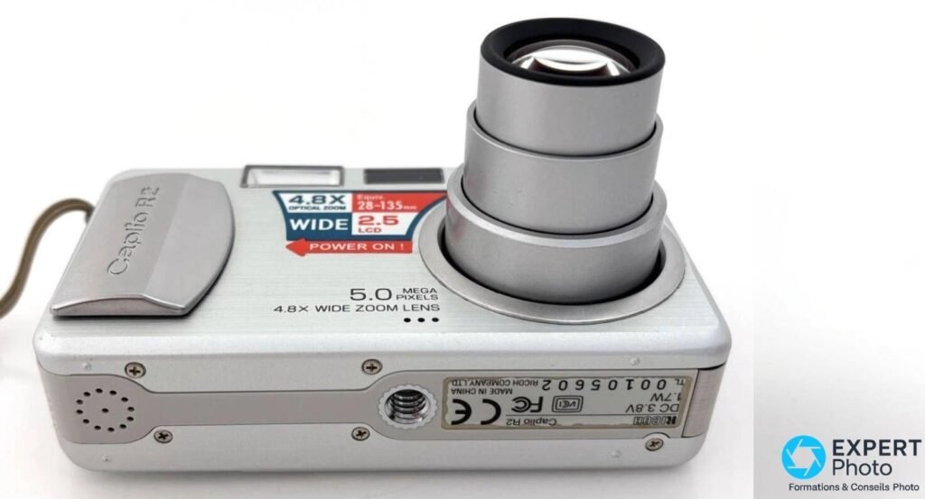 Ricoh Caplio R2 vu en 3/4 avec étiquette MADE IN CHINA RICOH COMPANY, filetage trépied 1/4 pouce et objectif déployé.