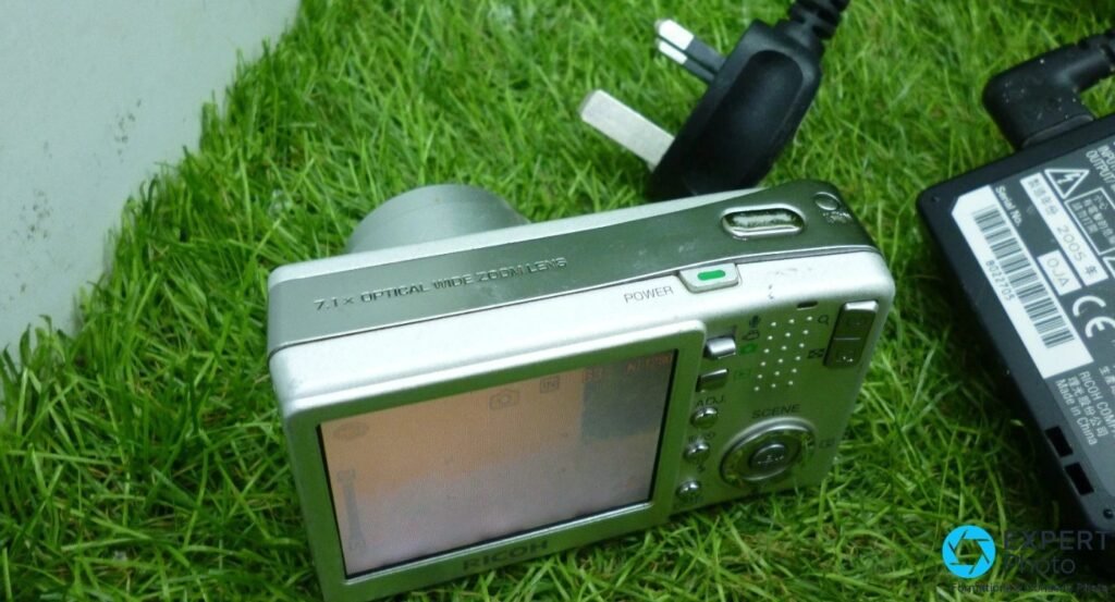 Vue arrière 3/4 du Ricoh Caplio R3 : écran LCD, boutons ADJ et SCENE, chargeur Ricoh BJ-6 visible.