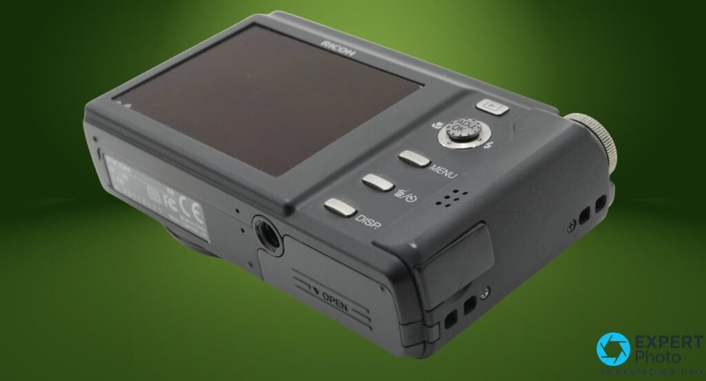 Ricoh Caplio R8 vu en 3/4 arrière en plongée, trappe batterie marquée OPEN, écrou trépied, étiquette réglementaire visible. 