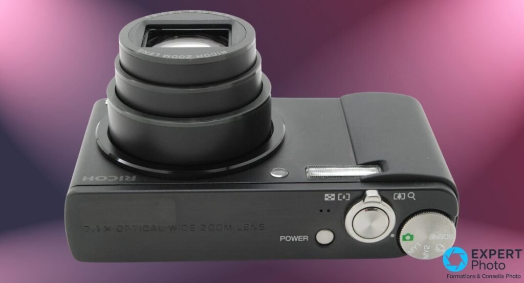 Ricoh Caplio R8 vu de dessus, molette de modes argentée avec icônes MY1 MY2 SCENE, bouton POWER, sur fond magenta. 