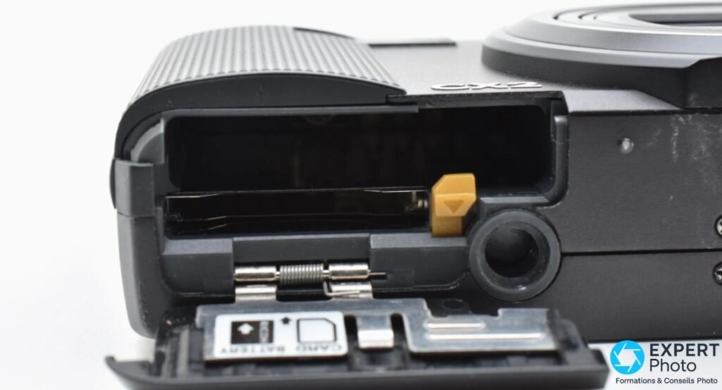 Trappe batterie et carte SD du Ricoh CX2 ouverte, avec ressort métallique et languette de blocage orange visibles.