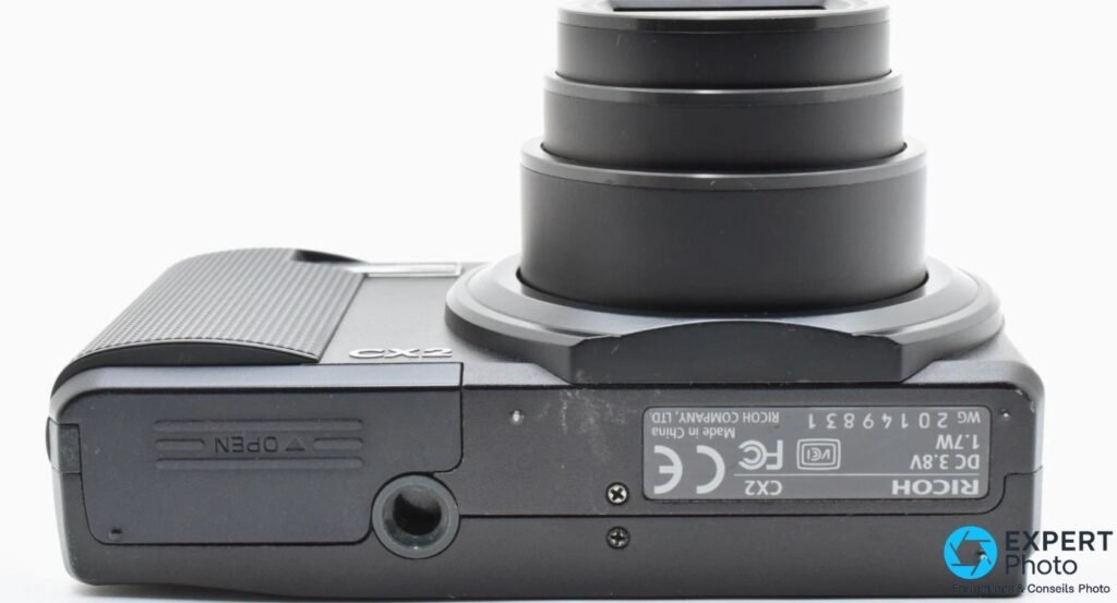 Vue de dessous du Ricoh CX2 montrant l'étiquette serial Ricoh, le pas de vis trépied et la trappe batterie fermée. 