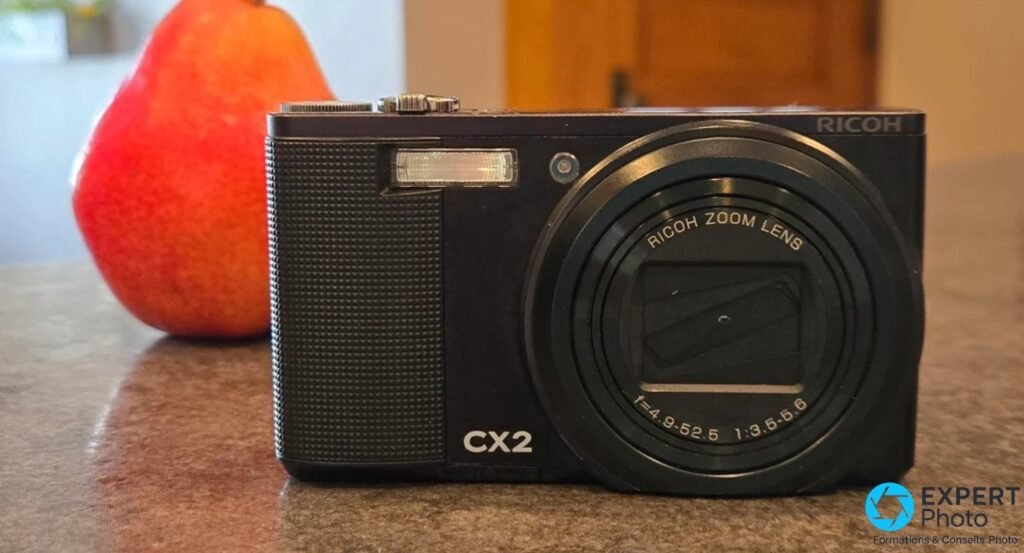 Ricoh CX2 noir vu de face, objectif rétracté, posé sur une table avec un fruit rouge flou en arrière-plan.