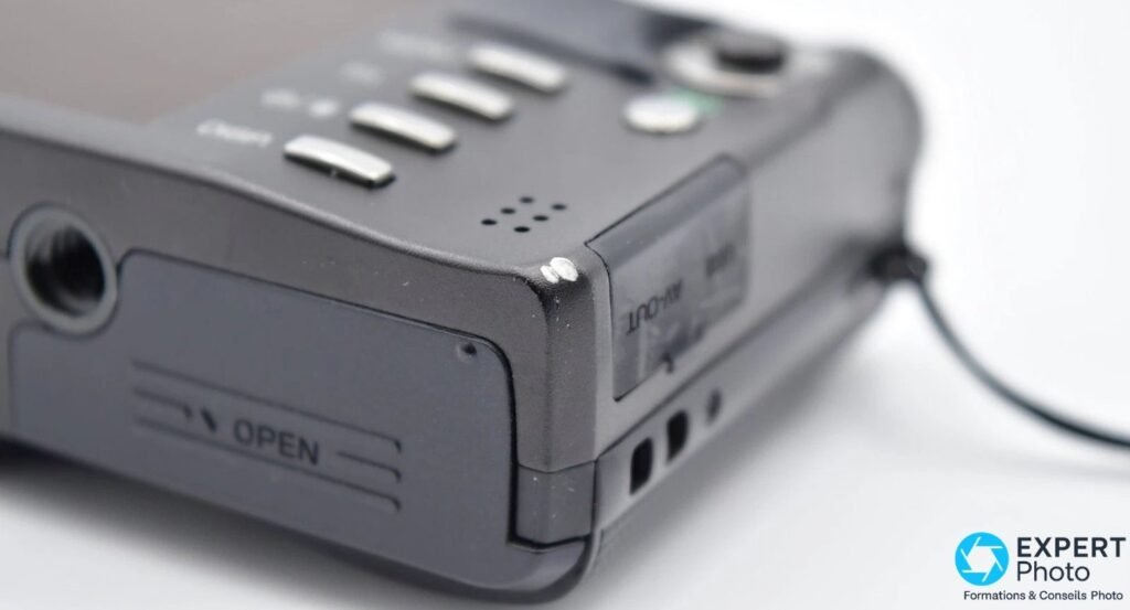 Détail Ricoh CX3 sur la trappe batterie marquée OPEN, pas de vis trépied visible et trappe AV-OUT à côté 